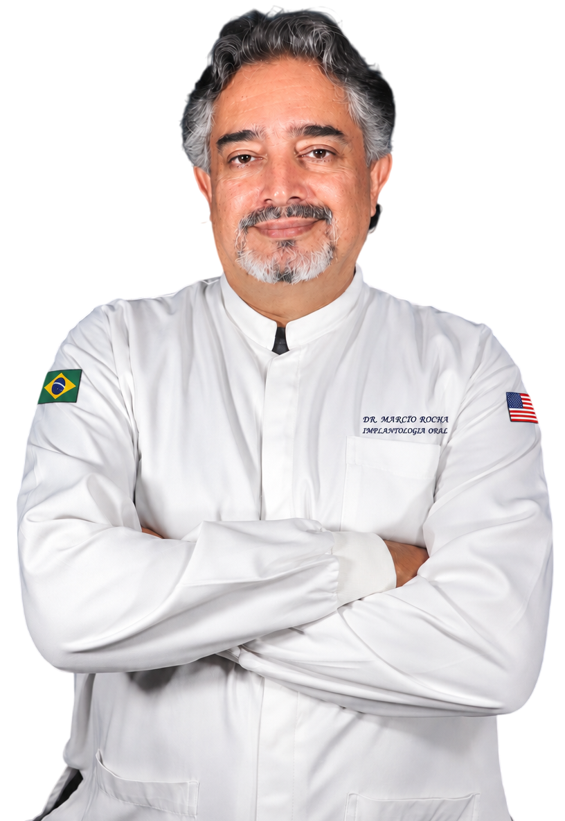 Médico de meia idade, sorrindo, com cabelos e barba grisalhos, usando um jaleco branco e de braços cruzados.
