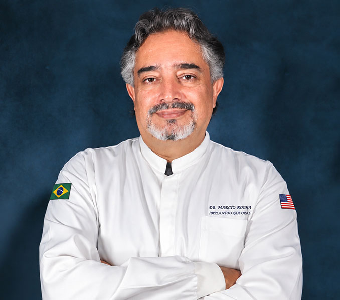 Médico de meia idade, sorrindo, com cabelos e barba grisalhos, usando um jaleco branco e de braços cruzados.